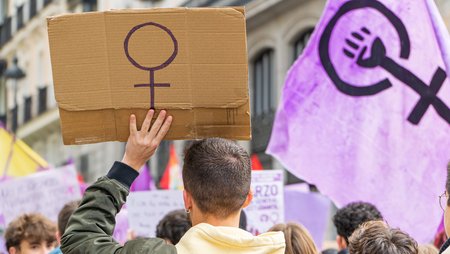 A man seen from behind holds a placard with the venus sign. He is joining a demonstration. Right in the picture is a purple flag showing the venus sign with a raised fist.