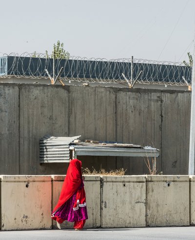 A woman with a red veil walk along a grey wall