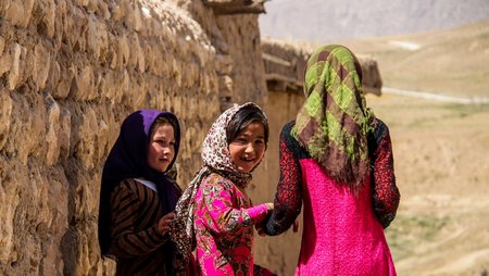 Drei junge afghanische Mädchen vor ländlicher Kulisse, zwei drehen sich um und blicken lächelnd in die Kamera.