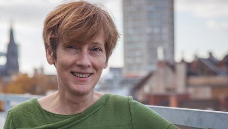 Eine Frau mit roten Haaren und grünem Pullover steht auf einer Dachterrasse mit Kölner Dom im Hintergrund. Es ist Martina Grantz, Bereichsleitung Fundraising bei medica mondiale.