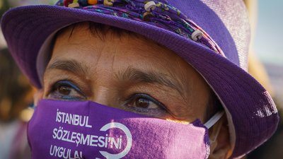 Nahaufnahme des Gesichts einer Frau mit lilafarbenem Hut und lilafarbenem Mundschutz mit der tükischen Aufschrift İstanbul Sözleşmesi uygula! Was auf Deutsch soviel wie Istanbul Konvention umsetzten heißt.