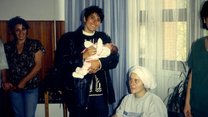 Eine Frau mit schwarzen Haaren hält in einem Zimmer ein Baby im Arm. 
