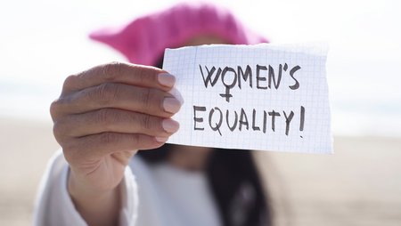 A woman holds a small handwritten sign towards the camera with the words “Women’s Equality!”