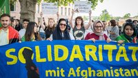 Viele Frauen und einige Männer stehen auf einem Platz. Sie halten ein großes, blaues Banner sowie kleinere Schilder. Auf einem Schild steht „In Solidarity With Afghan Women“.