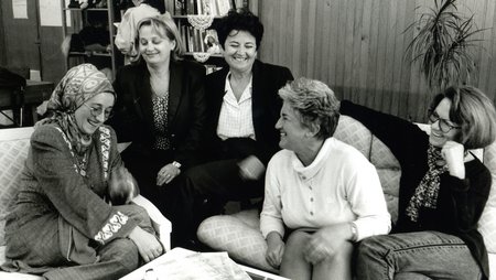 A group of women sit around a table, laughing. It is the team of the Medica Zenica women’s centre (black-and-white photo, 1997)
